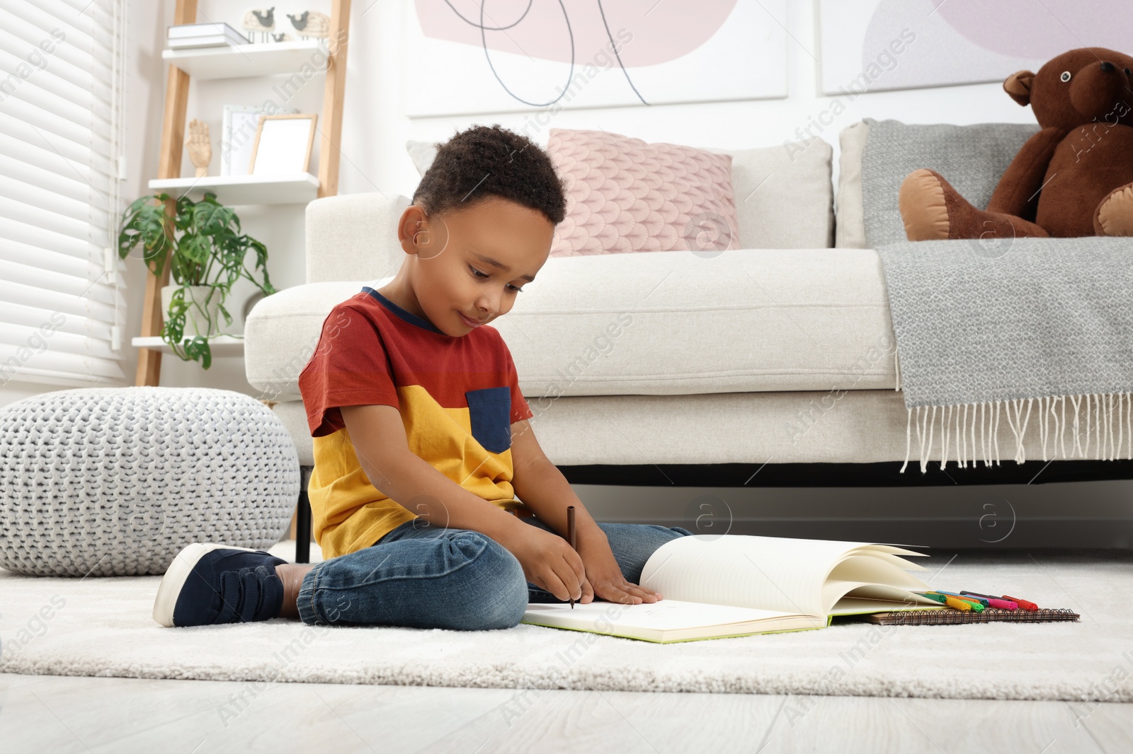 Photo of Cute African-American boy drawing in sketchbook with colorful markers on floor at home