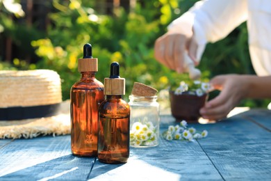 Photo of Woman grinding chamomile flowers in mortar outdoors, focus on bottles of essential oil