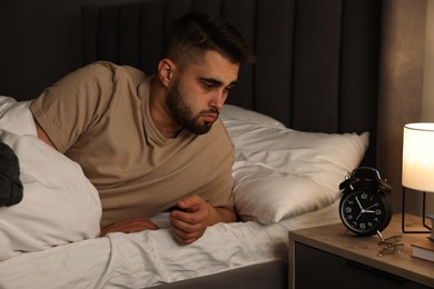 Photo of Frustrated man looking at alarm clock on bed