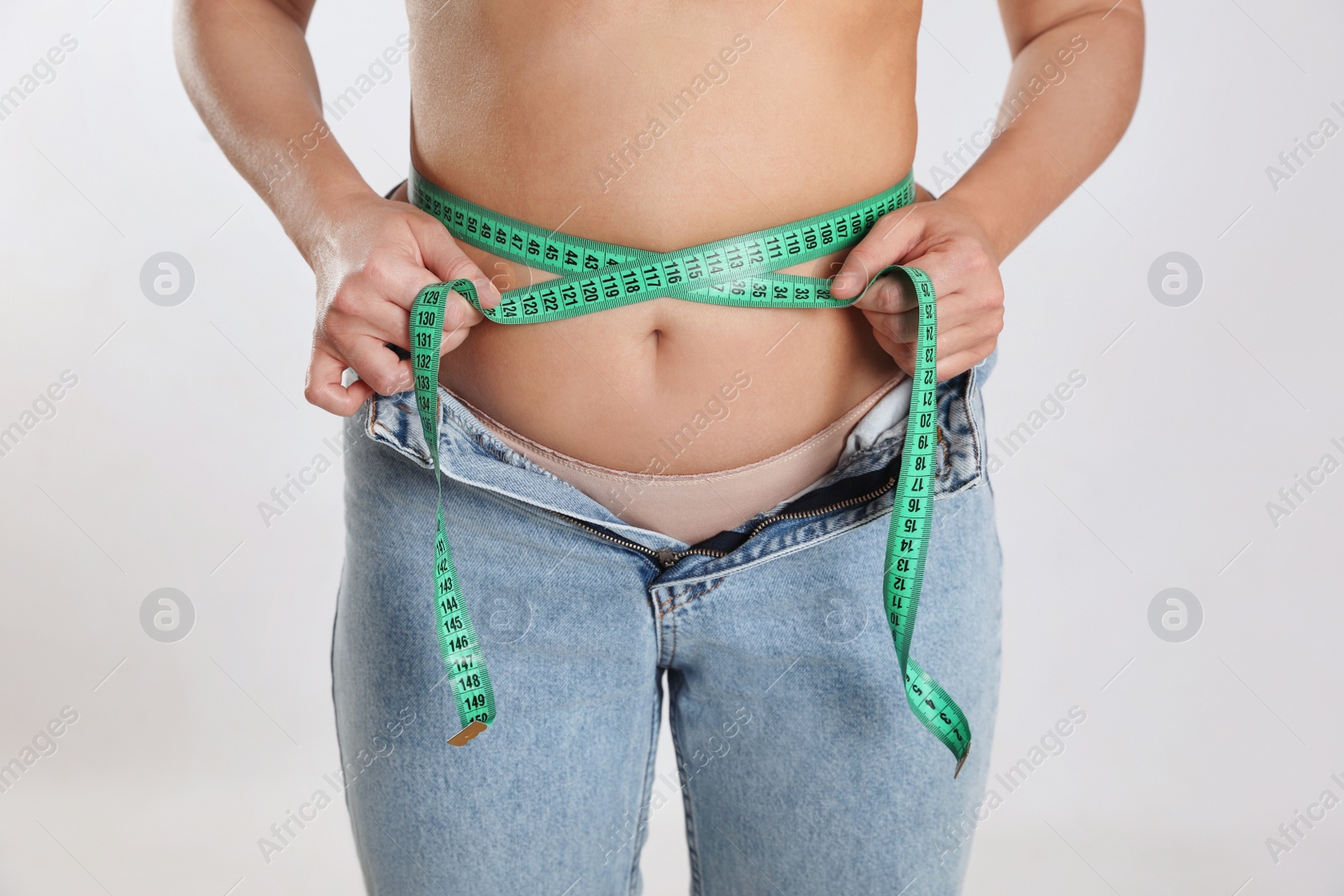 Photo of Woman in unfit jeans measuring her waist on light background, closeup. Weight loss concept