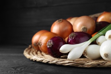 Wicker mat with different kinds of onions on black wooden table, closeup. Space for text