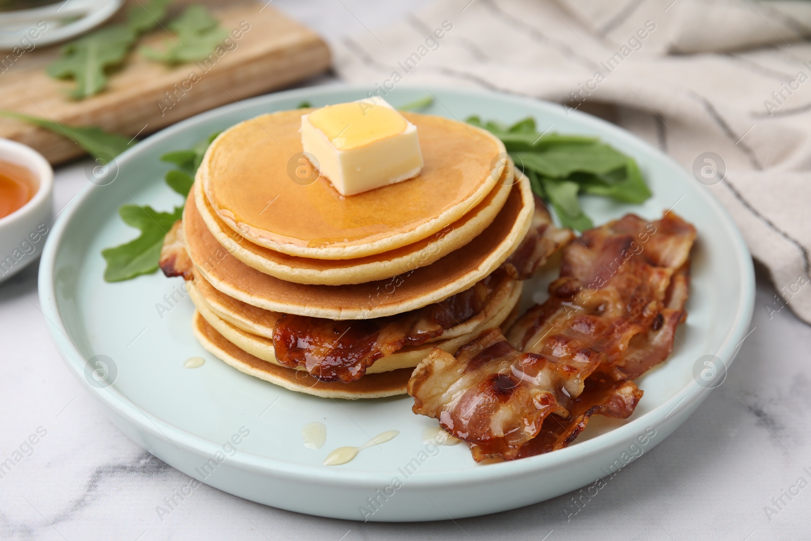 Photo of Tasty pancakes with butter, fried bacon and fresh arugula on white marble table