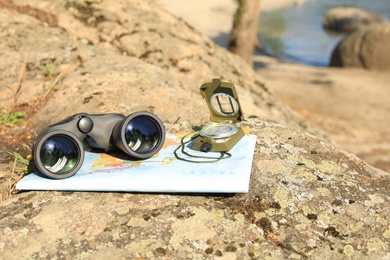 Binoculars, compass and map on stone near river, space for text. Camping equipment