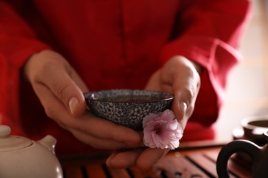 Photo of Master holding cup of freshly brewed tea and flower during traditional ceremony at table, closeup