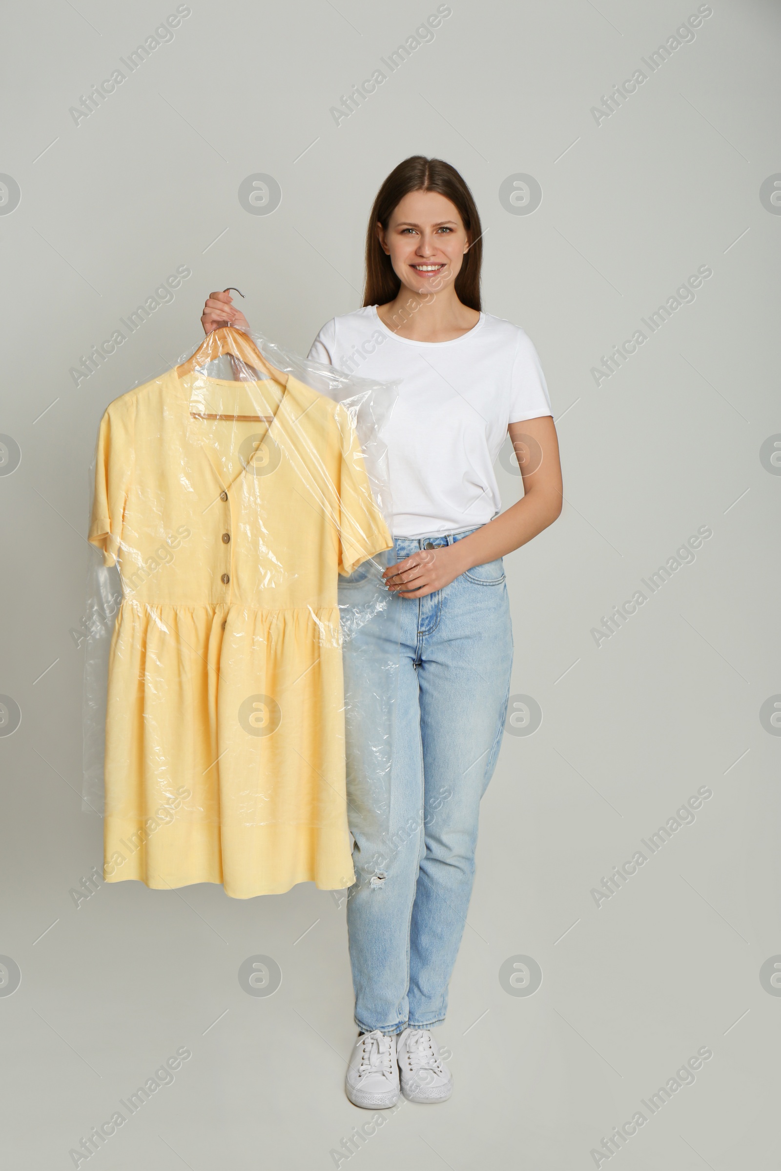 Photo of Young woman holding hanger with dress in plastic bag on light grey background. Dry-cleaning service