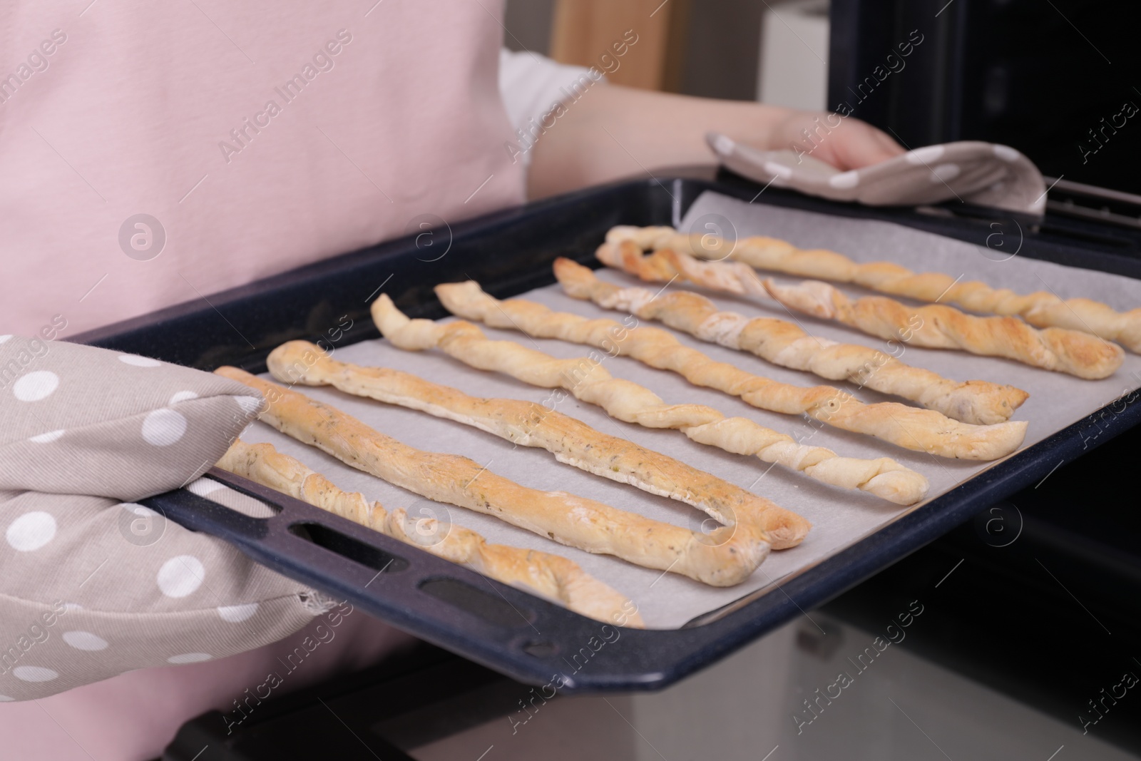 Photo of Woman holding baking sheet with homemade breadsticks near oven in kitchen, closeup. Cooking traditional grissini