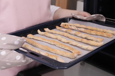 Woman holding baking sheet with homemade breadsticks near oven in kitchen, closeup. Cooking traditional grissini