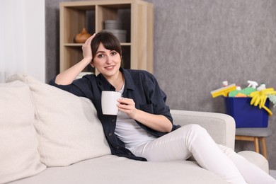 Beautiful young housewife with cup of drink resting after cleaning on sofa at home