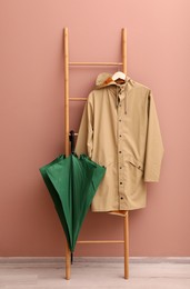 Photo of Stylish green umbrella and raincoat on wooden ladder near beige wall indoors