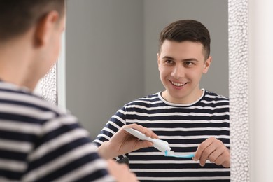 Happy man squeezing toothpaste from tube onto toothbrush near mirror in bathroom