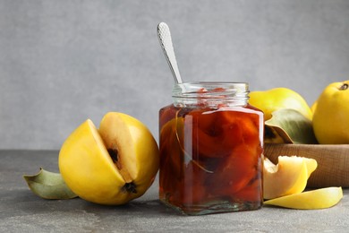 Photo of Tasty homemade quince jam in jar, spoon and fruits on grey textured table