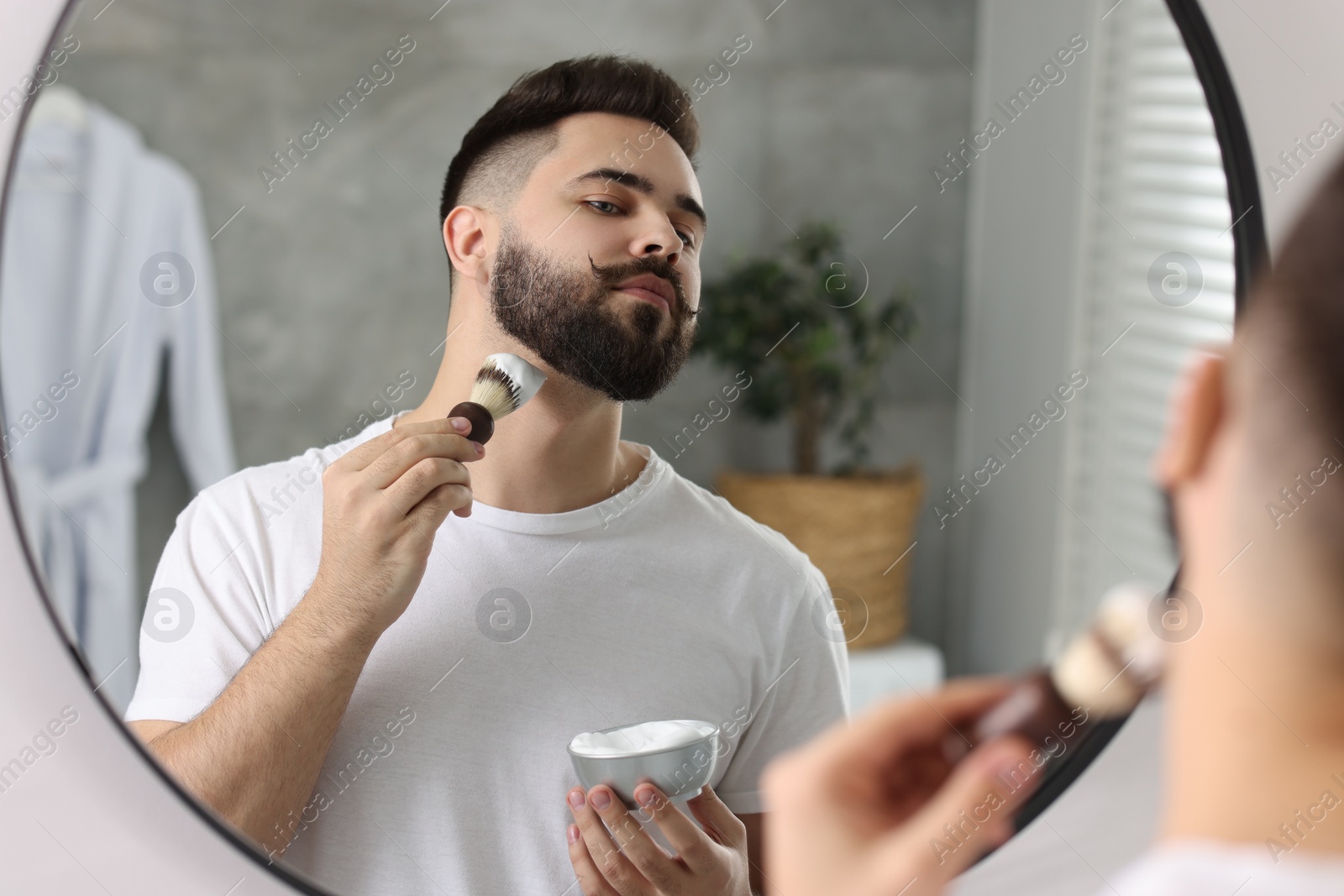 Photo of Handsome young man shaving beard near mirror in bathroom