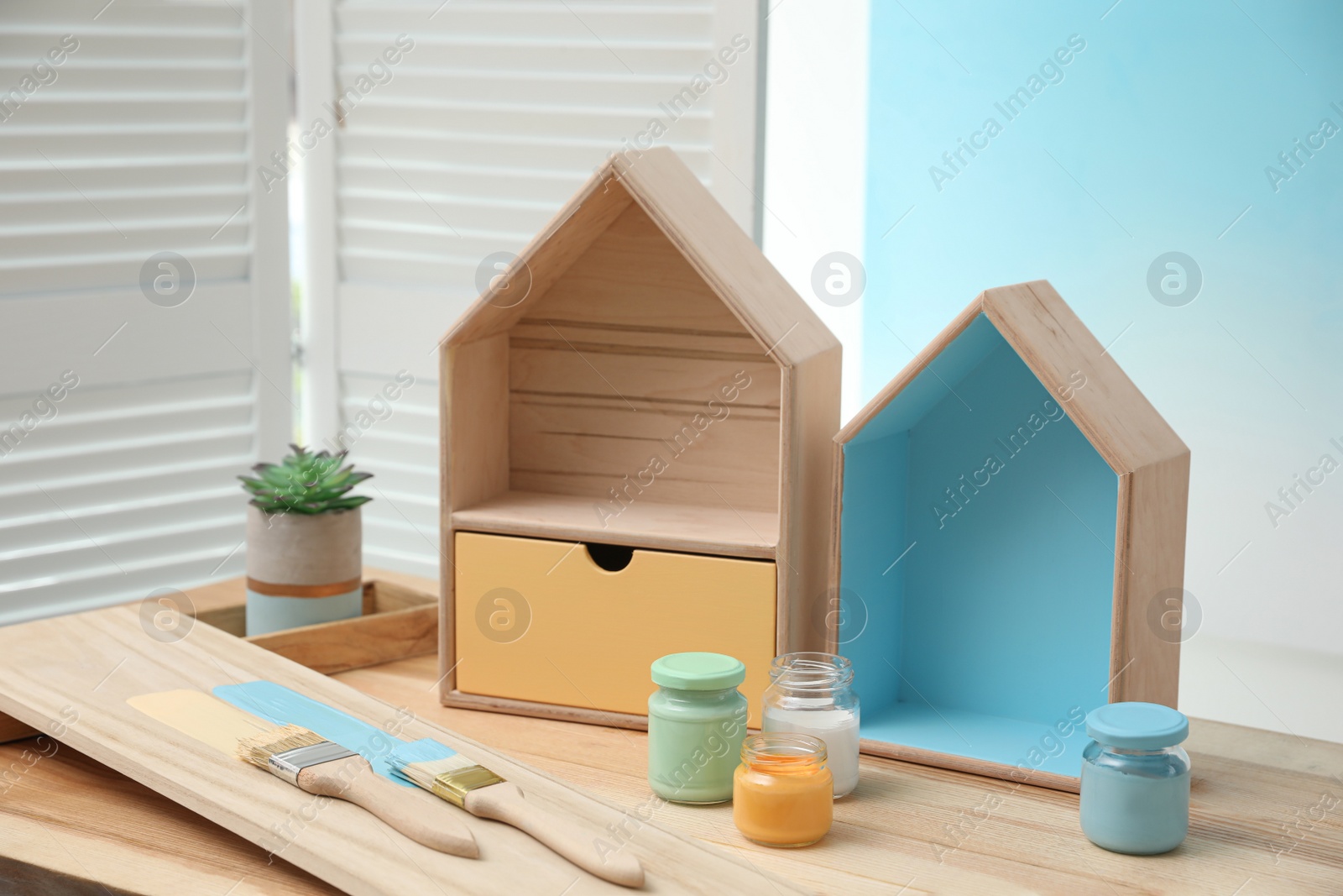 Photo of House shaped shelves, brushes and jars of paints on table indoors