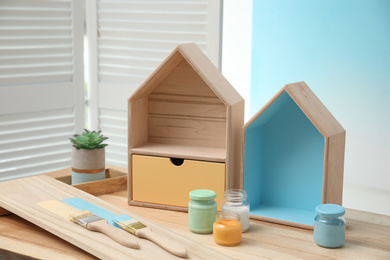 Photo of House shaped shelves, brushes and jars of paints on table indoors