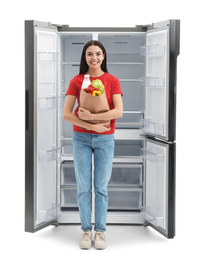 Young woman with bag of groceries near empty refrigerator on white background