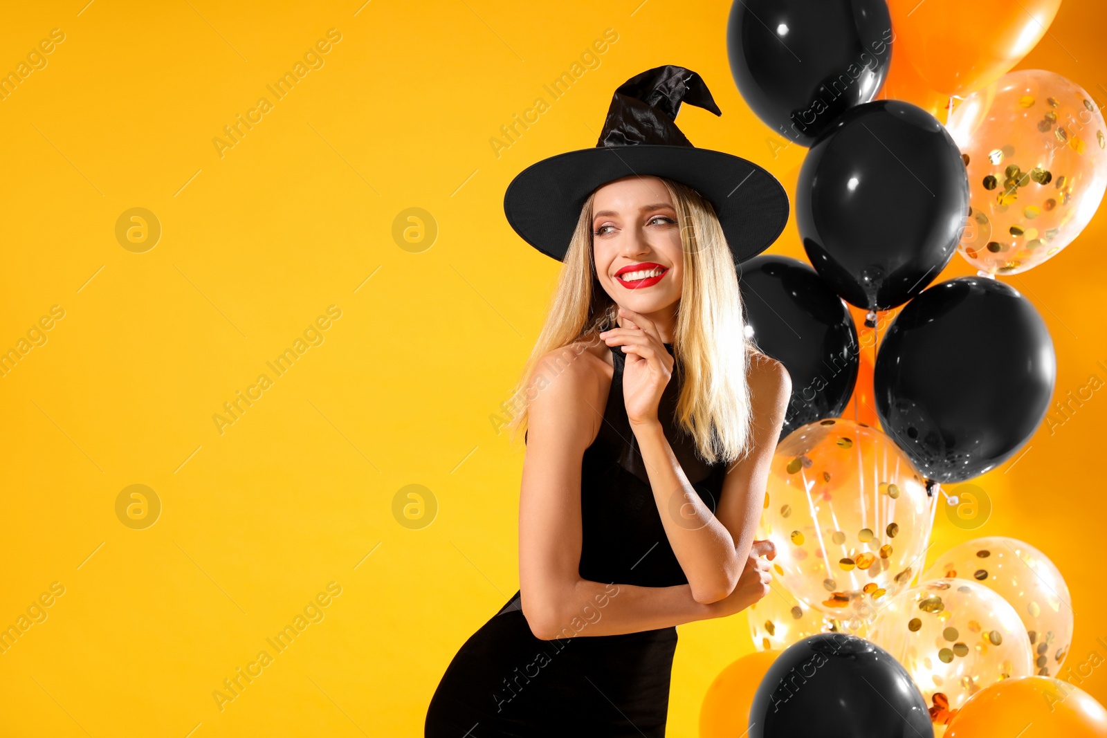 Photo of Beautiful woman in witch costume with balloons on yellow background, space for text. Halloween party