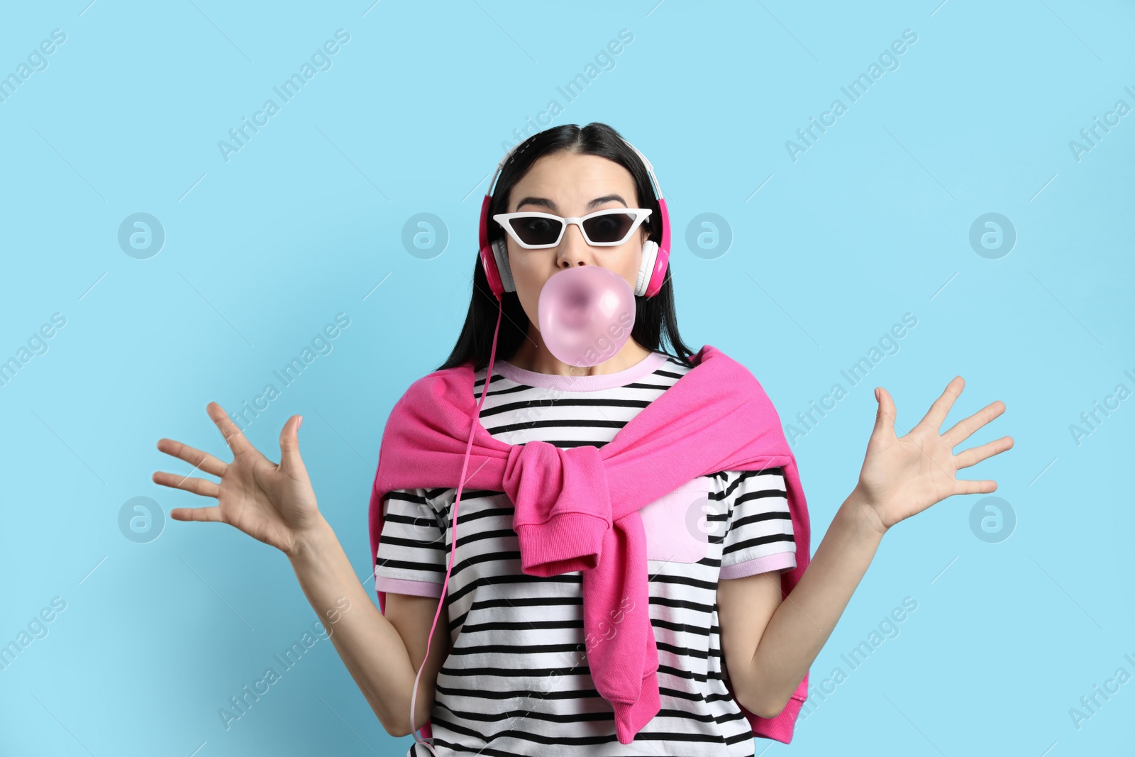 Photo of Fashionable young woman with headphones blowing bubblegum on light blue background