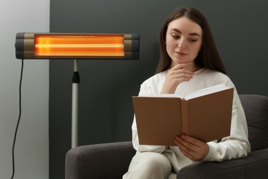 Young woman reading book on armchair near electric infrared heater at home. Space for text