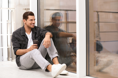 Photo of Handsome man with paper cup of coffee near window at home