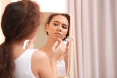 Beautiful young woman with acne problem looking in mirror at home