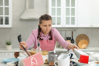 Photo of Upset woman in messy kitchen. Many dirty dishware and utensils on table