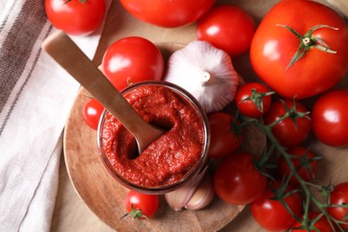 Jar of tasty tomato paste with spoon and ingredients on wooden table, flat lay