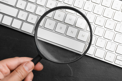 Woman holding magnifier glass over keyboard on black slate table, top view. Find keywords concept