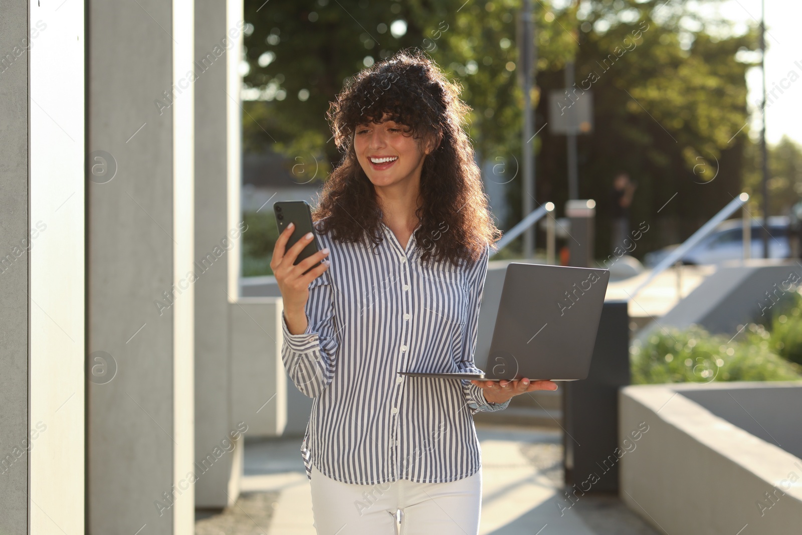 Photo of Happy young woman with modern laptop using smartphone outdoors