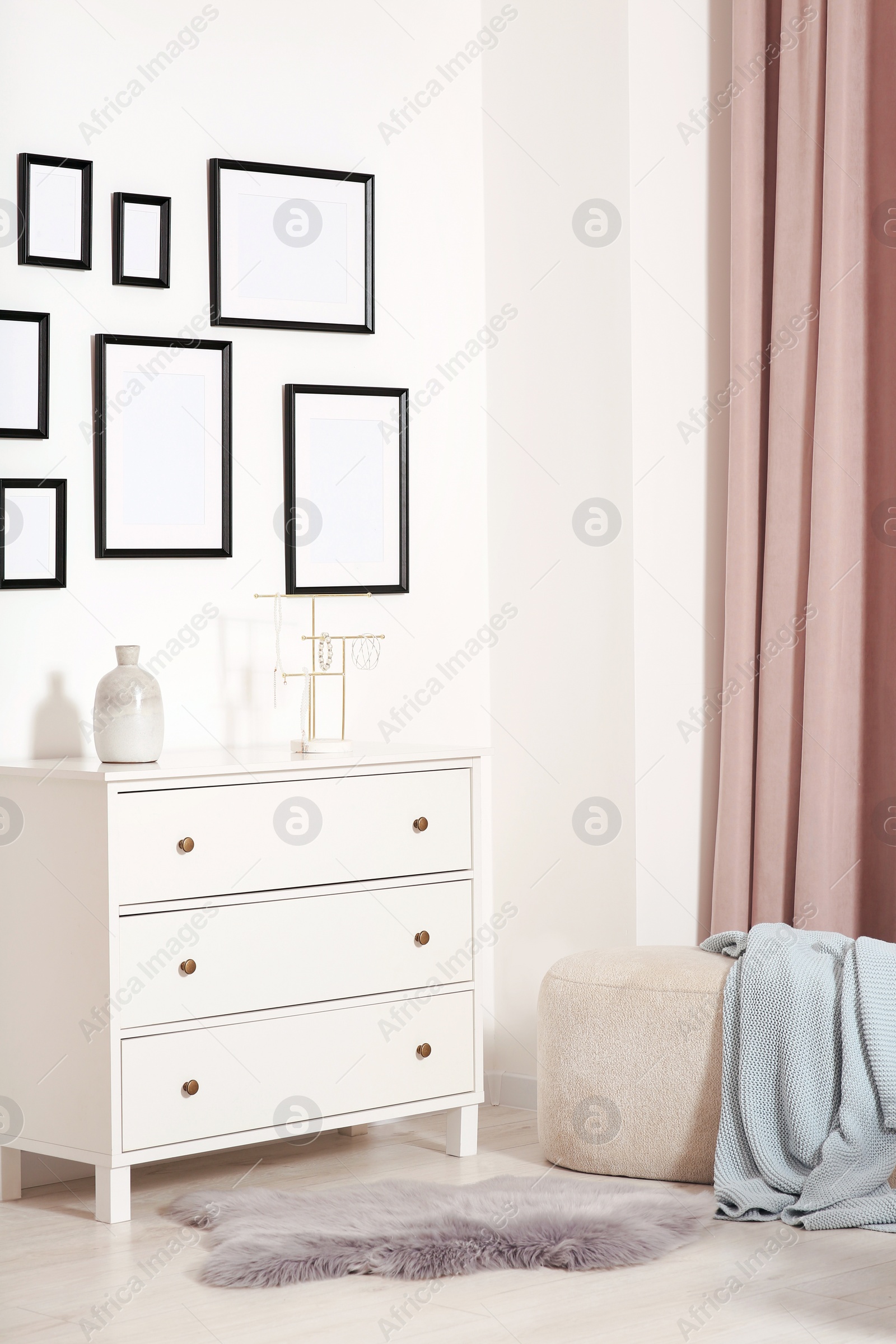 Photo of Empty frames hanging on white wall, chest of drawers and ottoman indoors