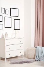 Photo of Empty frames hanging on white wall, chest of drawers and ottoman indoors