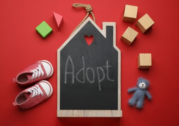 Photo of Small chalkboard with word ADOPT, toy bear, cubes and baby shoes on red background, flat lay