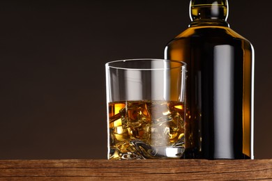 Whiskey with ice cubes in glass and bottle on wooden table, closeup. Space for text