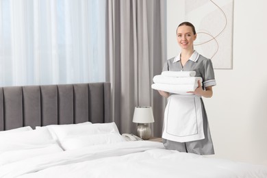 Photo of Young maid with stack of towels near bed in hotel room. Space for text