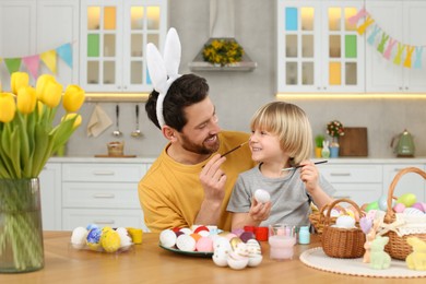Father and his cute son having fun while painting Easter eggs at table in kitchen