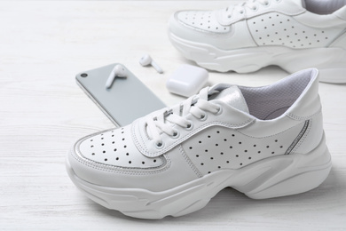 Photo of Pair of stylish sneakers, modern smartphone and wireless earphones on white wooden table