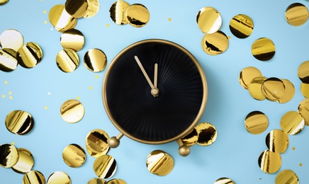 Alarm clock and golden confetti on light blue background, flat lay. New Year countdown