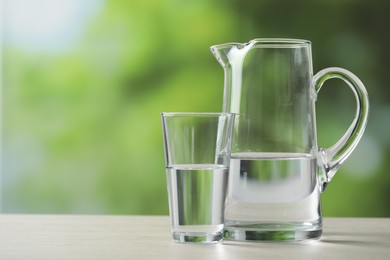 Photo of Jug and glass with clear water on white table against blurred green background, closeup. Space for text