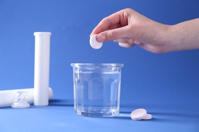 Woman putting effervescent pill into glass of water on blue background, closeup