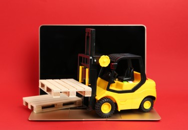 Laptop, toy forklift and wooden pallets on red background