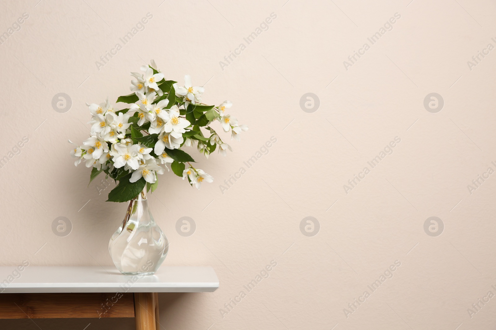 Photo of Bouquet of beautiful jasmine flowers in glass vase on table near beige wall, space for text