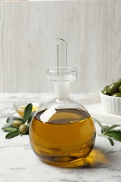 Photo of Glass jug of oil, ripe olives and green leaves on white marble table