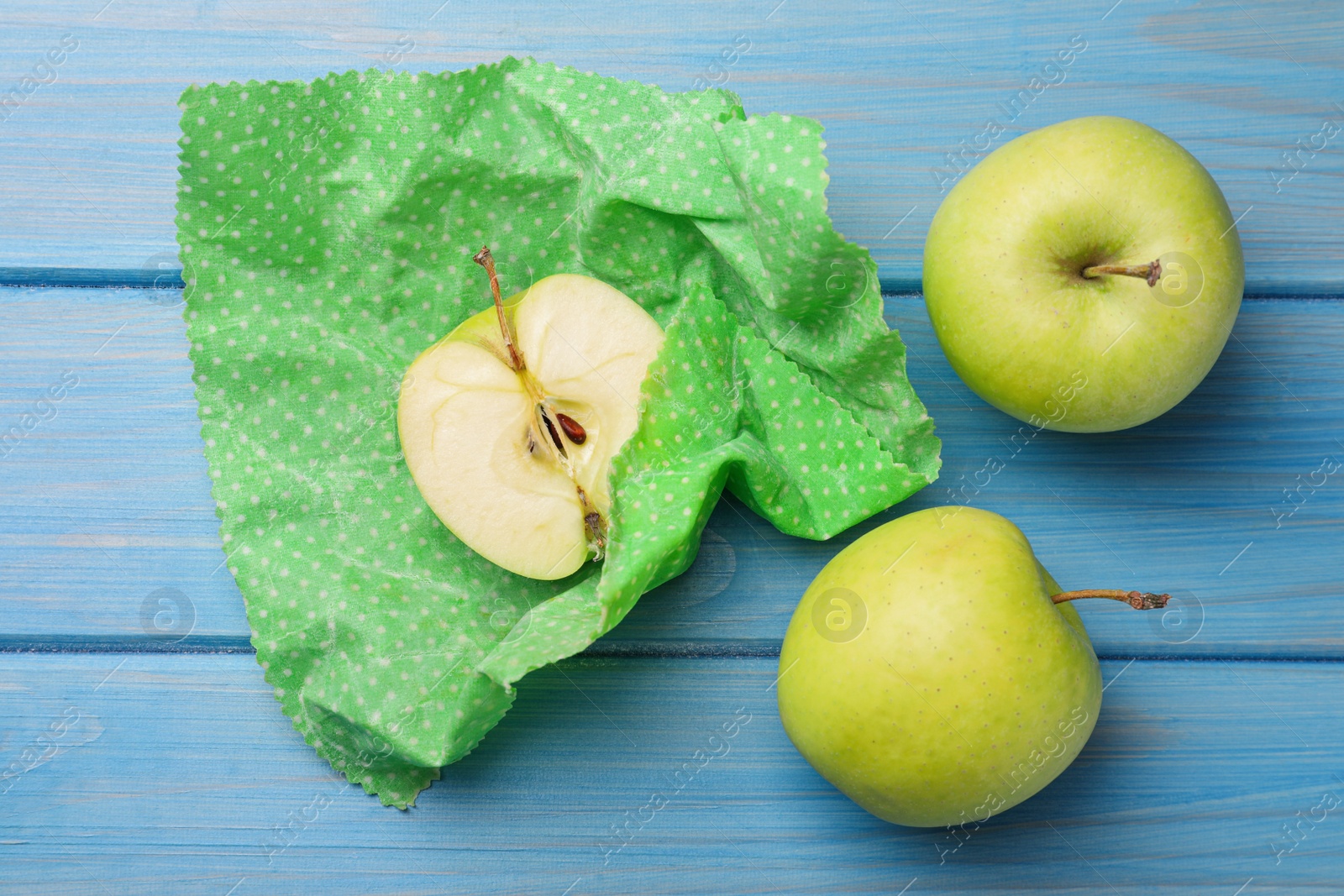 Photo of Whole and cut apples with beeswax food wrap on light blue wooden table, flat lay