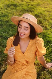 Photo of Beautiful young woman eating ice cream glazed in chocolate on green grass outdoors