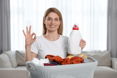 Photo of Woman holding fabric softener near basket with dirty clothes and showing OK gesture in room