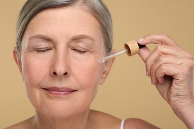 Senior woman applying cosmetic product on her aging skin against beige background. Rejuvenation treatment
