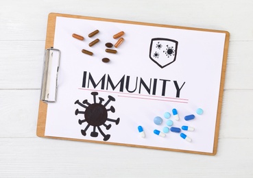 Clipboard with word Immunity and pills on white wooden table, top view