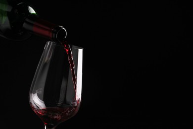 Pouring tasty red wine from bottle into glass on black background, closeup. Space for text