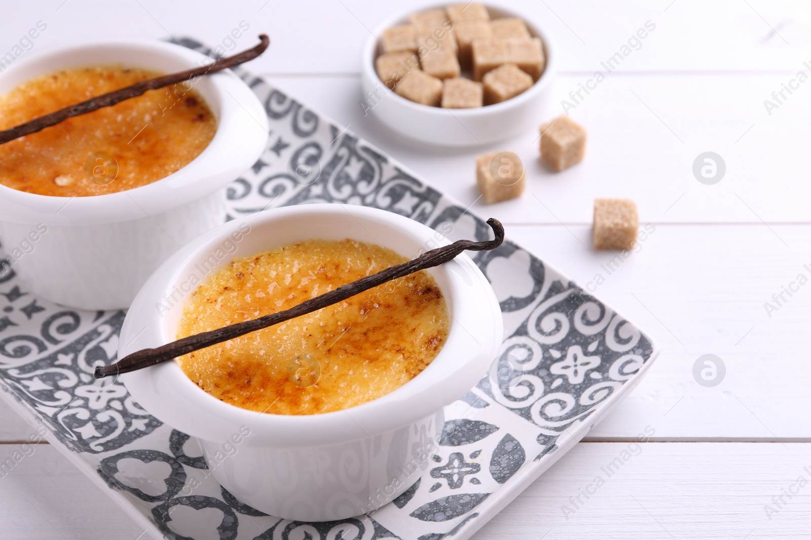 Photo of Delicious creme brulee in bowls, vanilla pods and sugar cubes on white wooden table, closeup