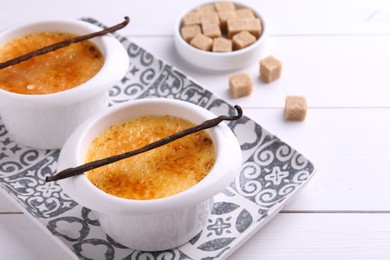 Photo of Delicious creme brulee in bowls, vanilla pods and sugar cubes on white wooden table, closeup