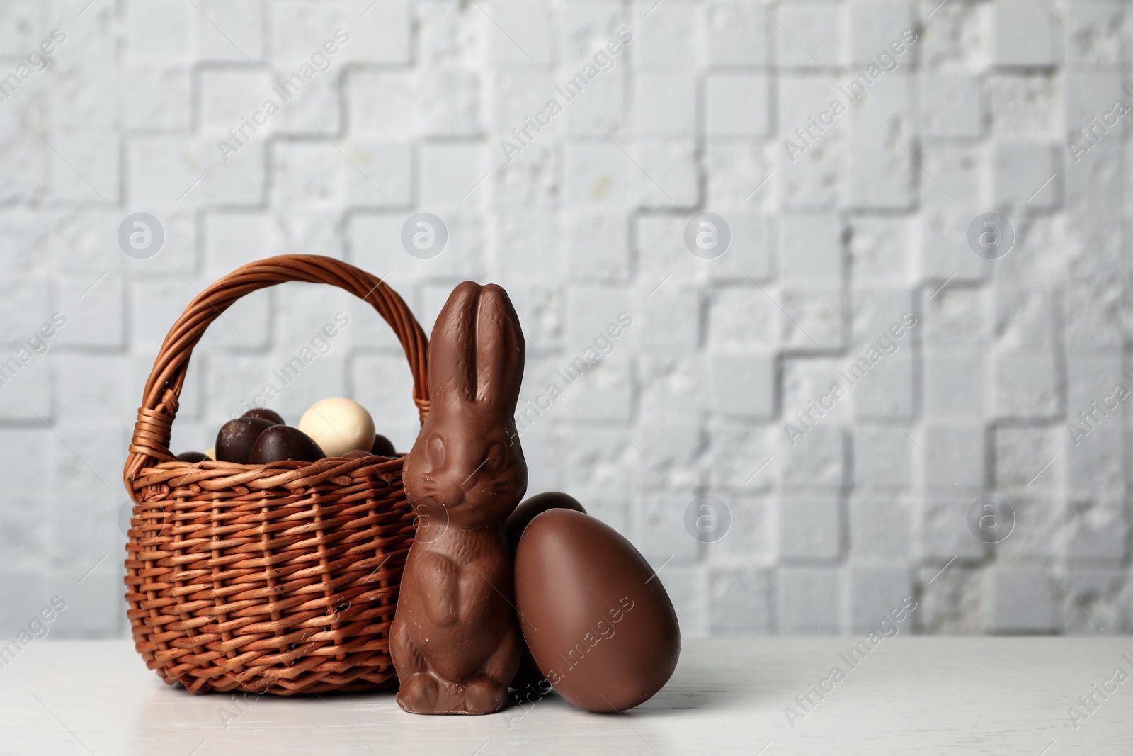 Photo of Wicker basket with sweet chocolate Easter eggs and bunny on table against light background, space for text